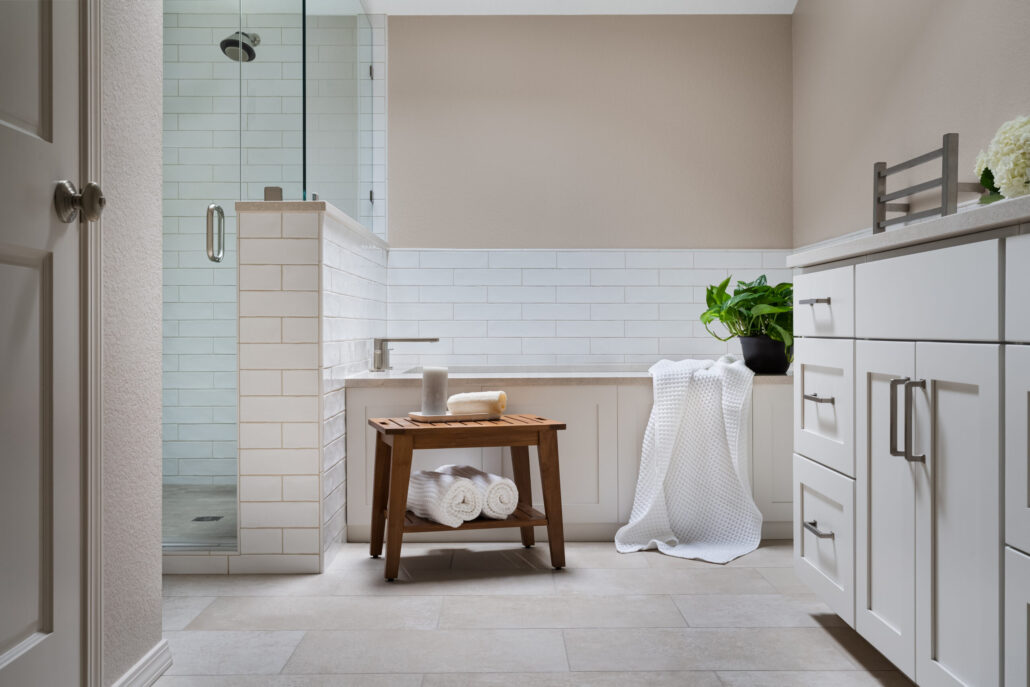 Modern bathroom with glass shower, white tiles, wooden bench, and plant decor. Fresh and clean design.