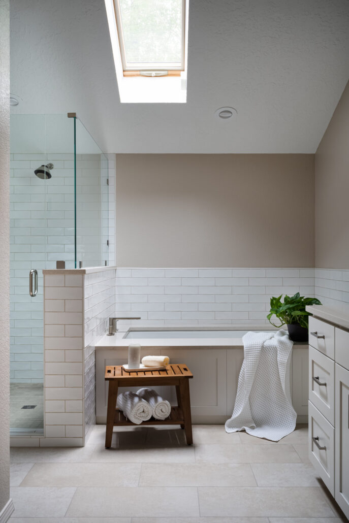 Modern bathroom with skylight, glass shower, and tub with plant. Cozy towels on wood stool for a spa-like feel.