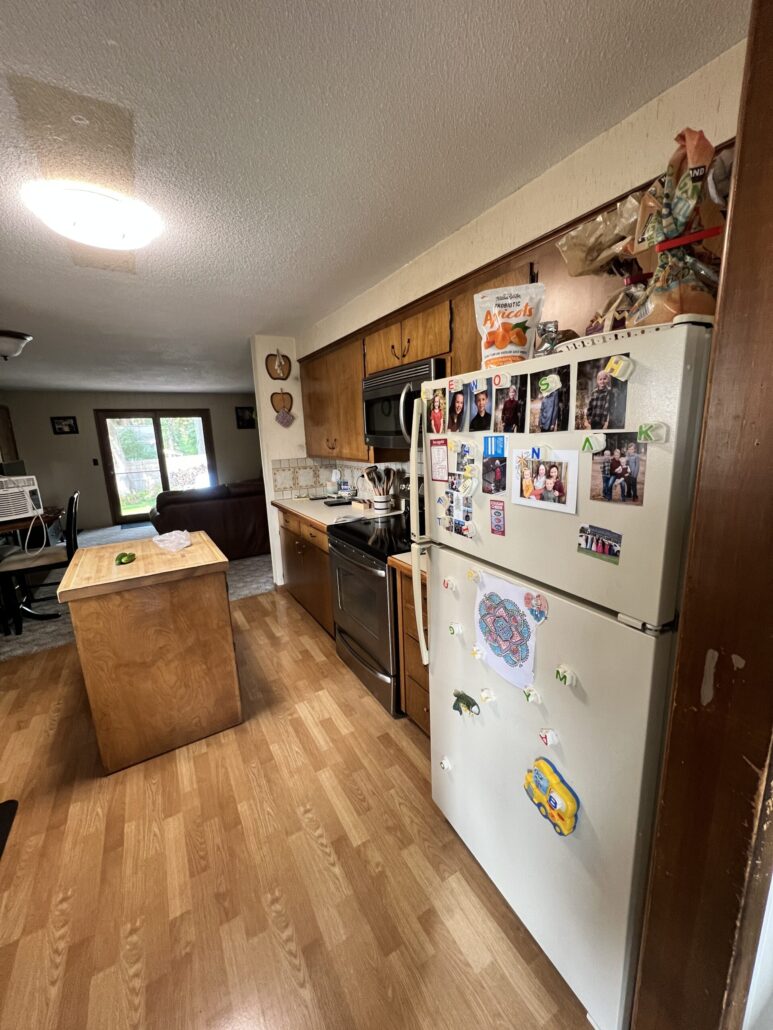 Cozy kitchen with wooden cabinets, island, and fridge covered in photos. Bright natural light from window.