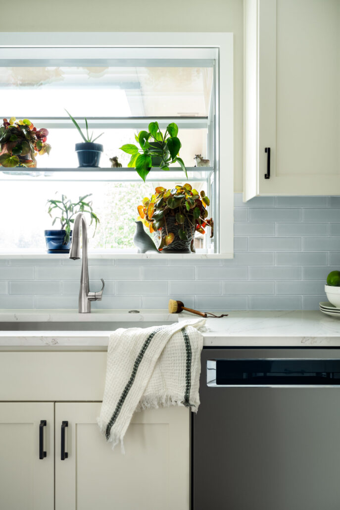 Modern kitchen sink with green plants on the windowsill, light gray tile backsplash, and white cabinets.