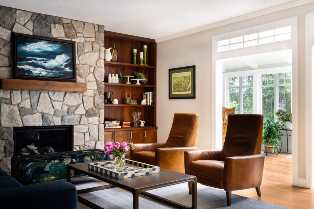 Cozy living room with leather chairs, stone fireplace, wooden shelves, and natural light from large windows.