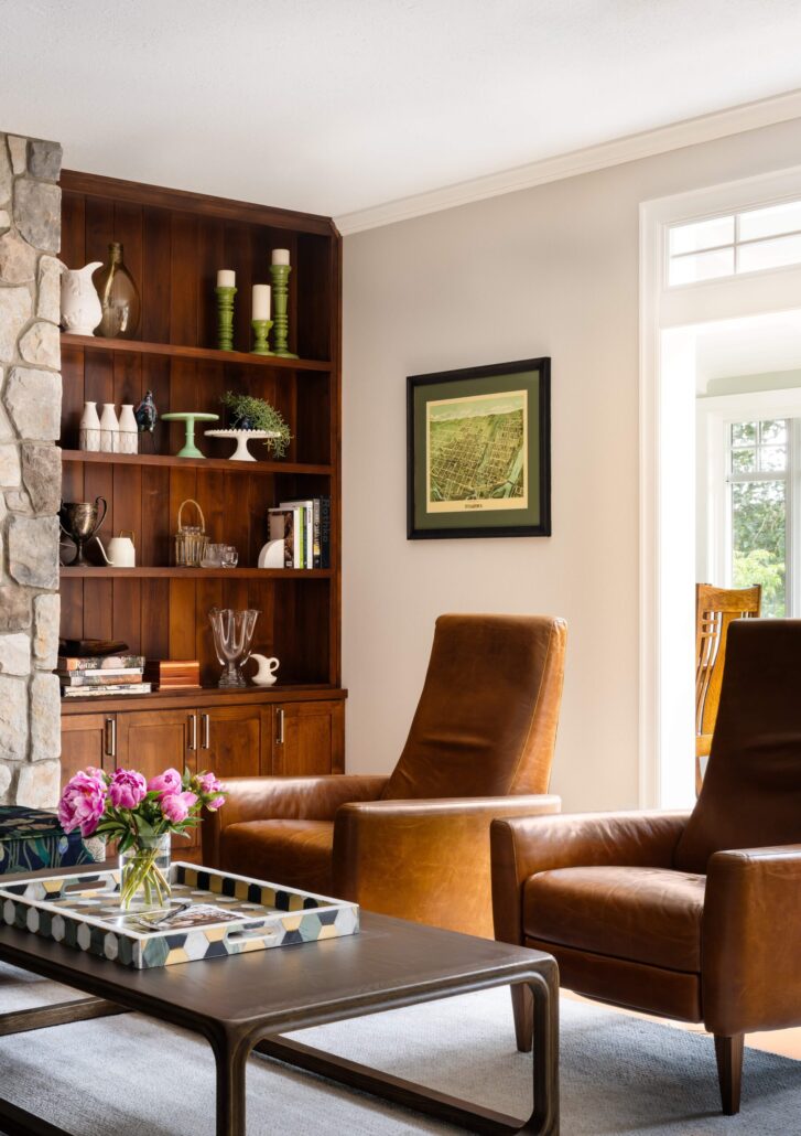 Cozy living room with leather chairs, wooden shelves, stone fireplace, and floral centerpiece on a coffee table.