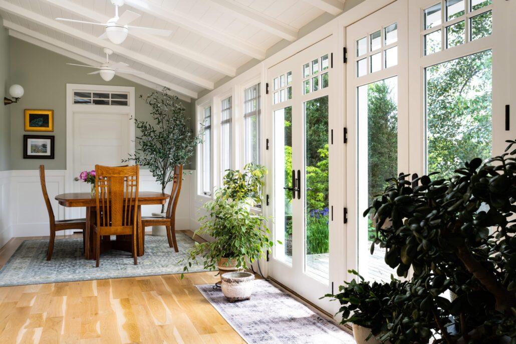 Bright dining room with wooden table, green plants, and large glass doors opening to a lush garden view.