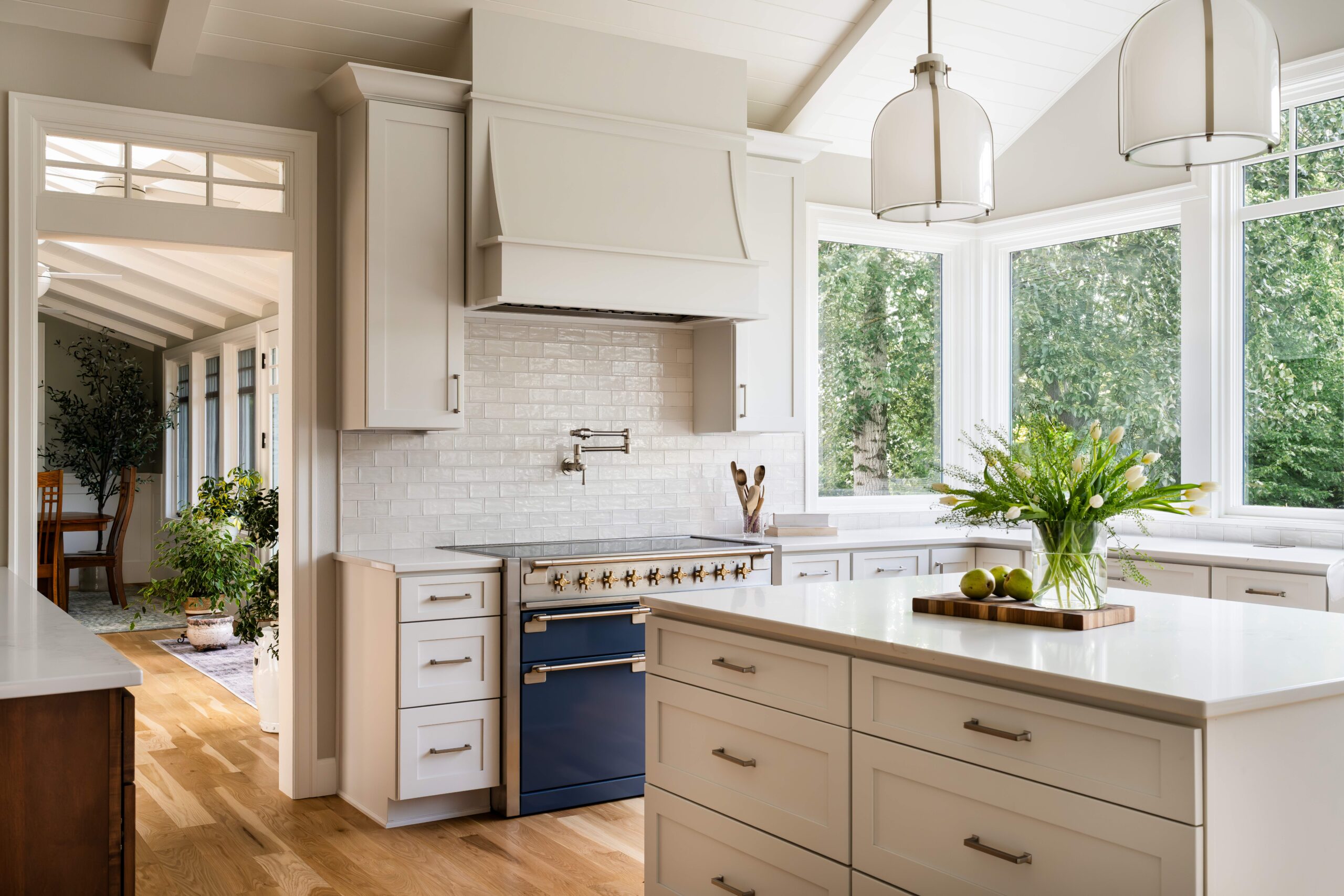 Bright, modern kitchen with white cabinets, blue oven, and large windows overlooking greenery. Airy and elegant design.