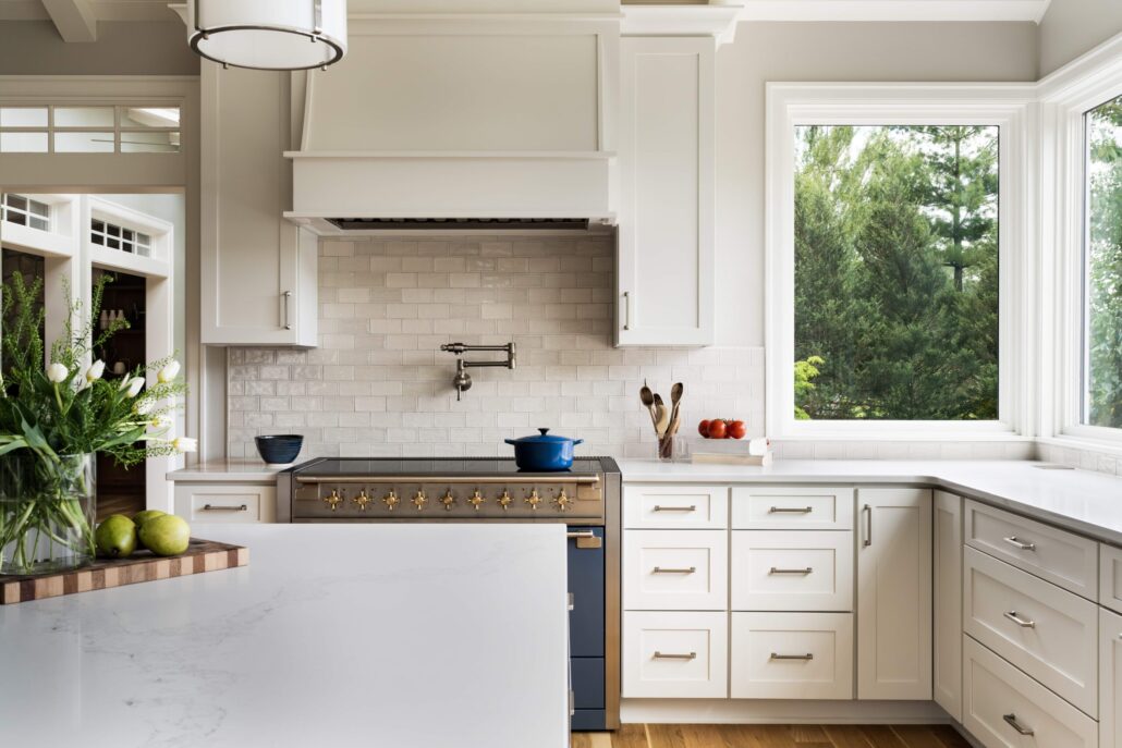 Modern kitchen with white cabinets, large window, and blue pot on stove. Elegant countertop with green apples and flowers.
