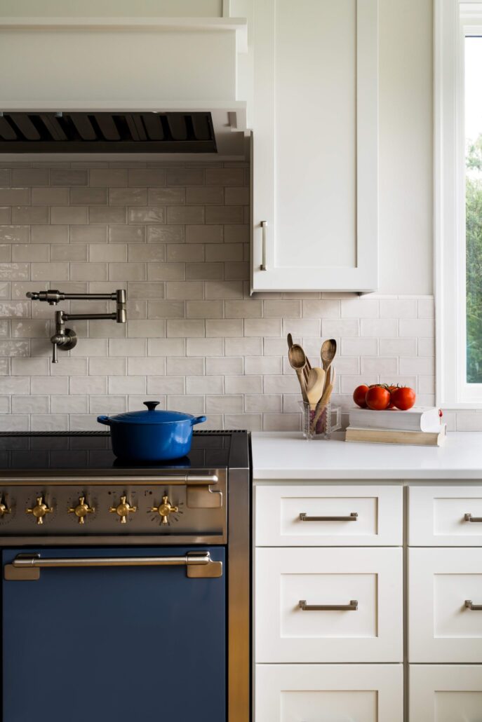 Modern kitchen with blue stovetop, white cabinets, and cooking utensils. Tomatoes and books on counter. Bright and clean.