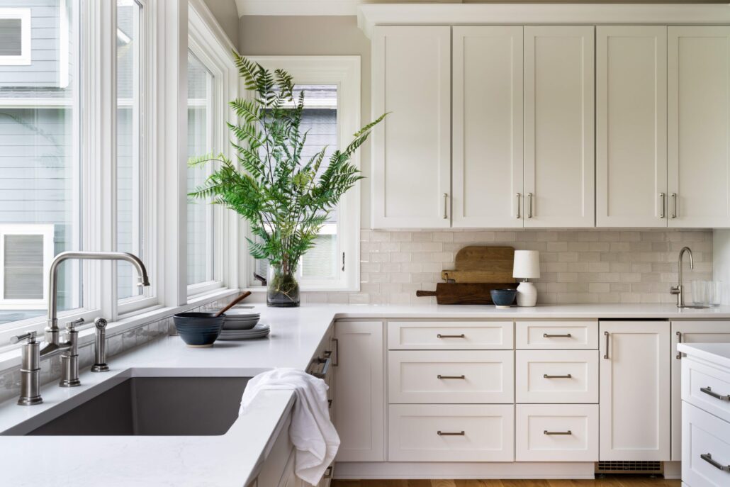 Modern kitchen with white cabinets, large windows, stainless steel sink, and green plant for a fresh, airy look.