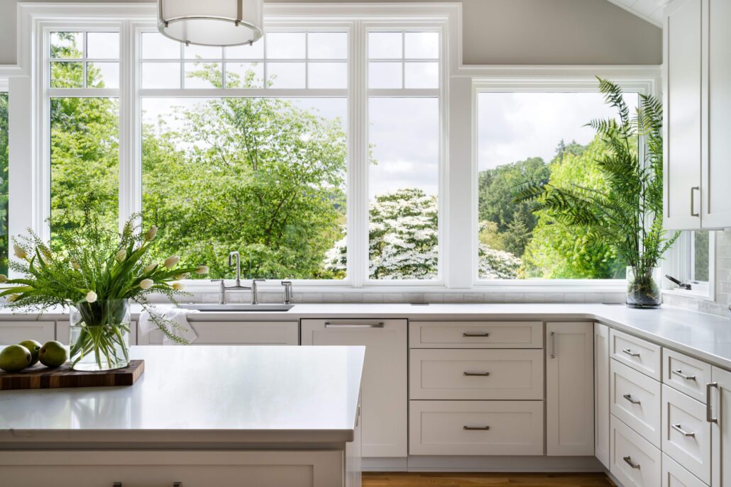 Modern white kitchen with large windows, lush greenery, and fresh flowers on the countertop, bright and airy design.