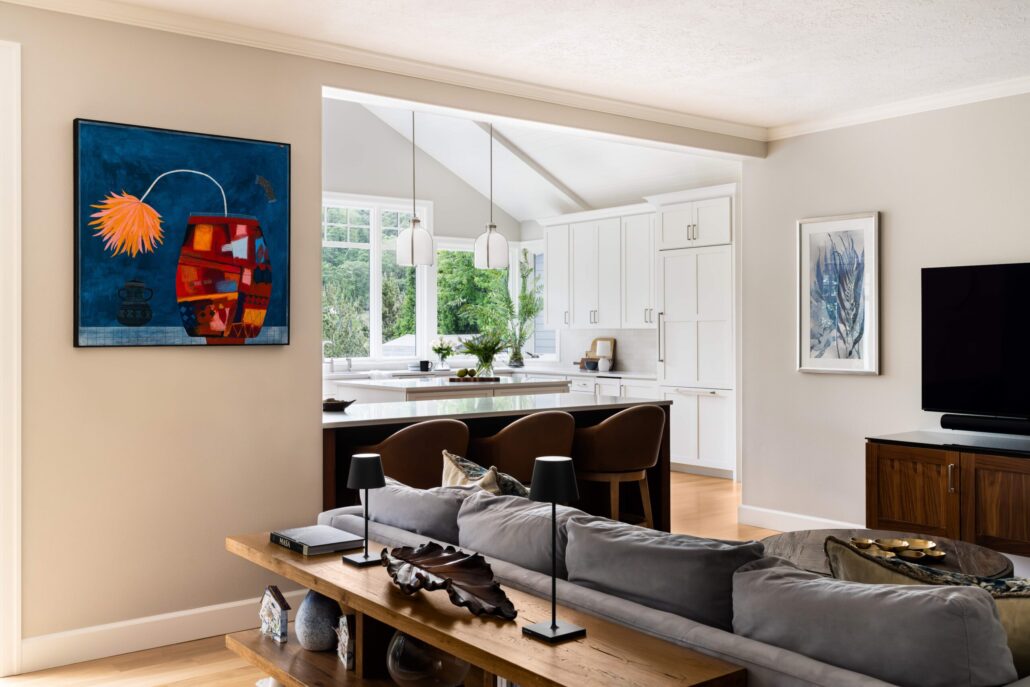 Modern living room with abstract art, view into bright kitchen with white cabinets and pendant lights.