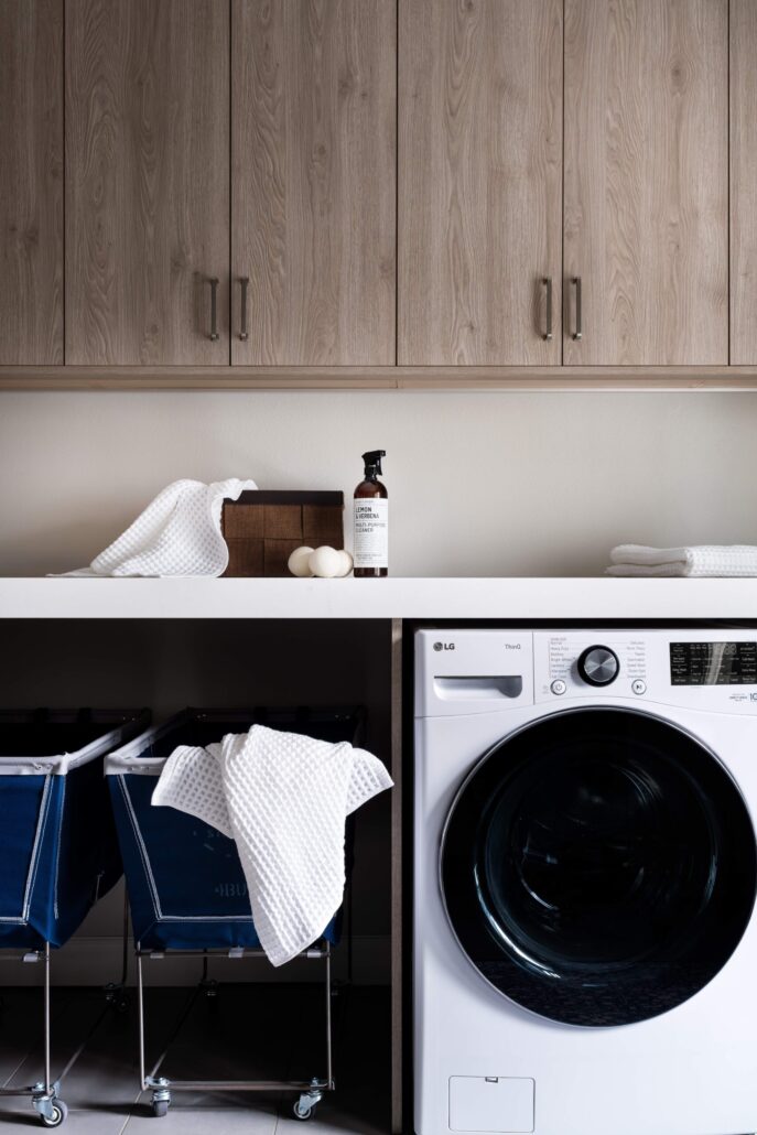 Modern laundry room with washer, cabinets, baskets, and cleaning products on a white countertop.