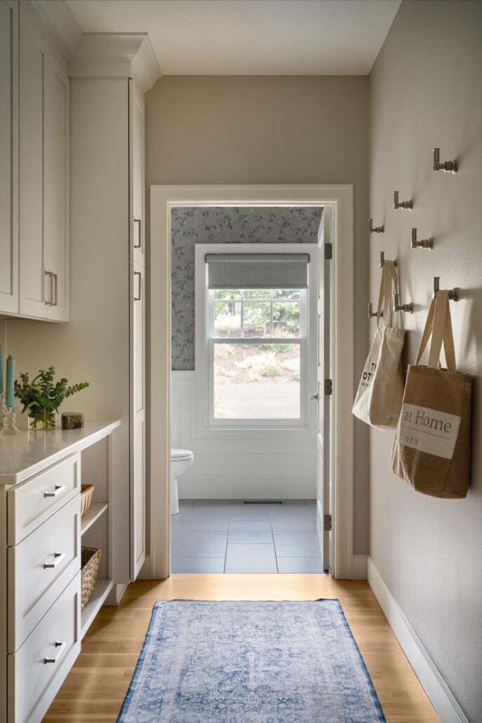 Bright hallway leading to a bathroom with blue rug, white storage, and wall hooks with bags hanging.