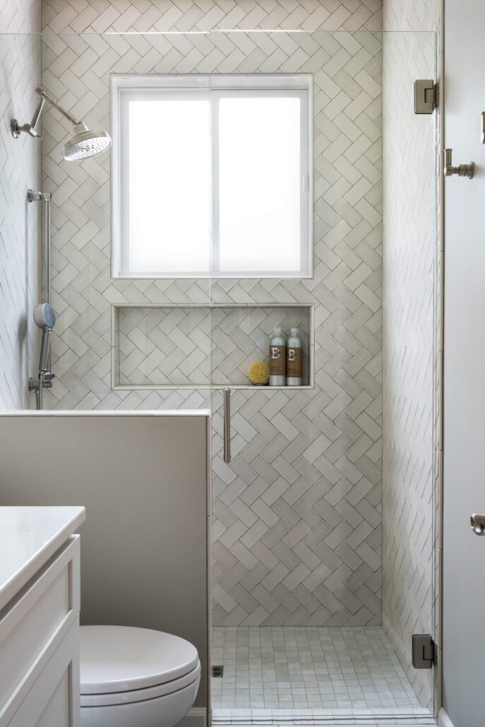 Modern bathroom with herringbone tile shower, rain showerhead, glass door, and built-in niche storage.