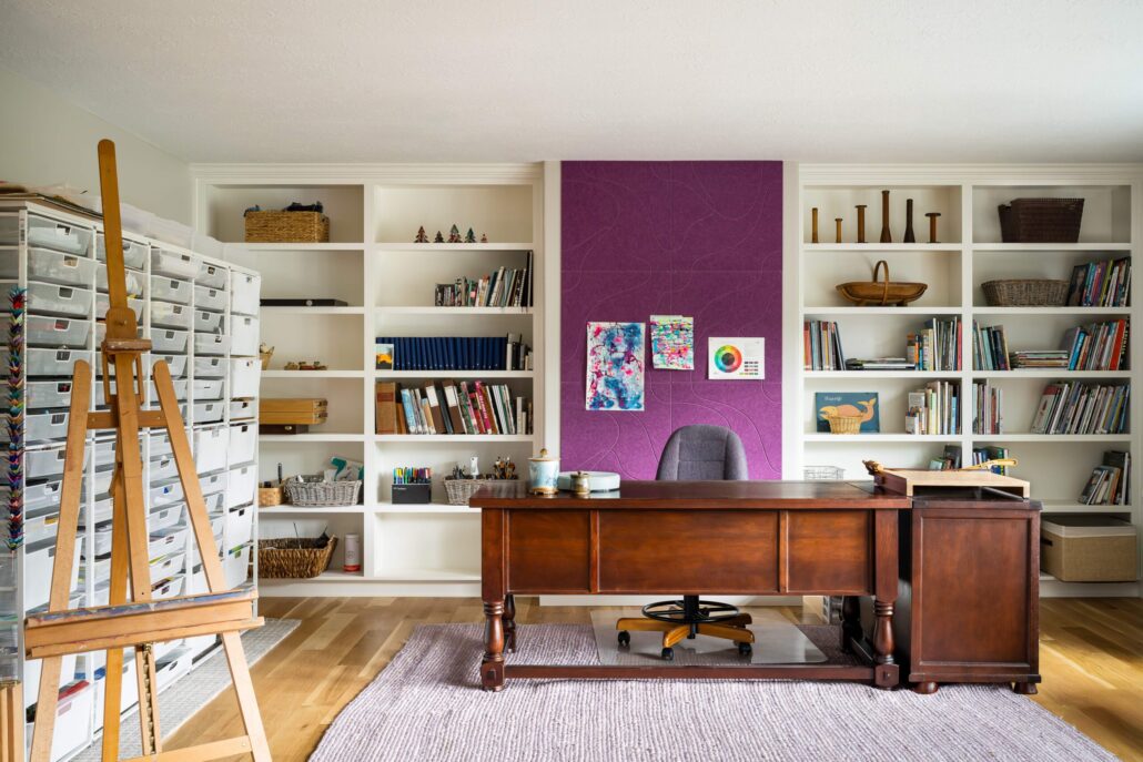 Creative home office with wooden desk, purple accent wall, bookshelves, and art supplies on an easel.