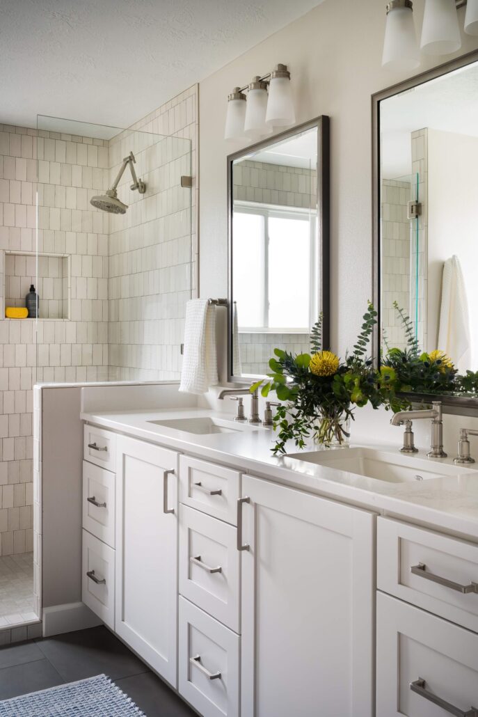 Modern bathroom with dual sinks, white cabinetry, and a glass shower with tile wall. Fresh greenery adds a pop of color.