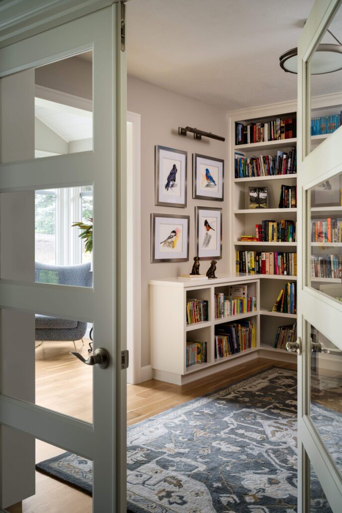Cozy home library with bird art, bookshelves, and decorative rug. Open door leading to a sunlit seating area.
