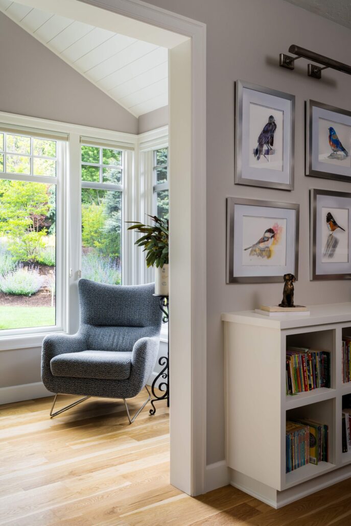 Cozy room with a gray armchair by a window, framed bird art, and bookshelves on wooden floor.