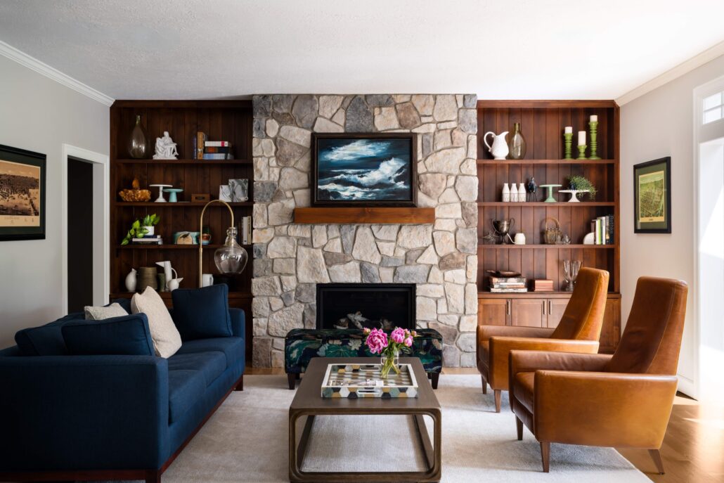 Cozy living room with stone fireplace, wooden shelves, blue sofa, brown chairs, and coffee table with flowers.