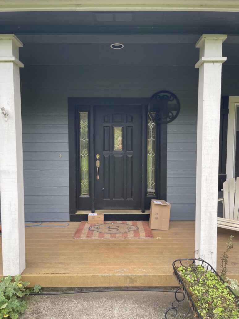 Front porch with a black door and two packages on a welcome mat, surrounded by white columns and a plant bed.
