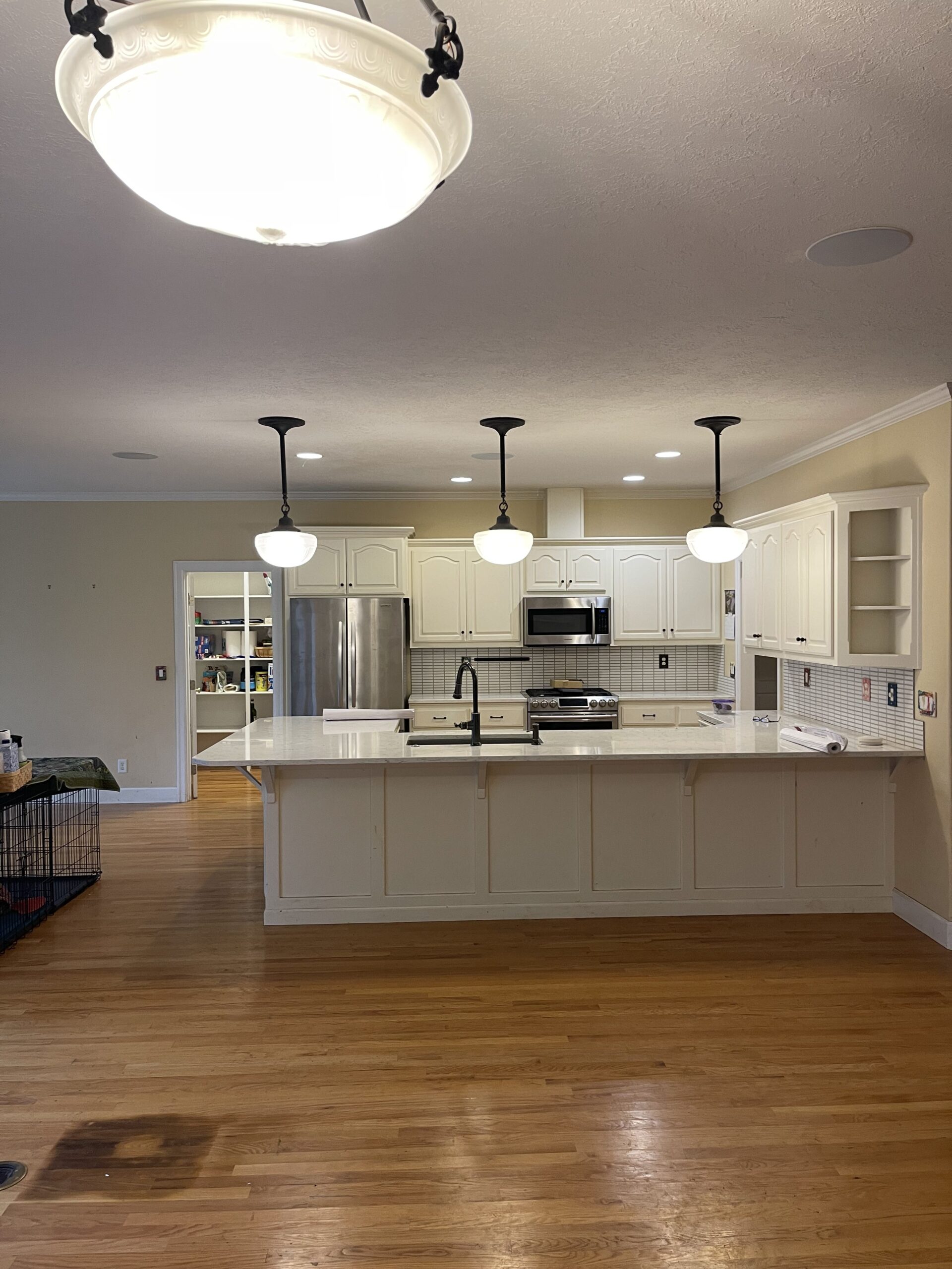 Old kitchen with white cabinetry, island, pendant lights, stainless steel appliances, and hardwood flooring.