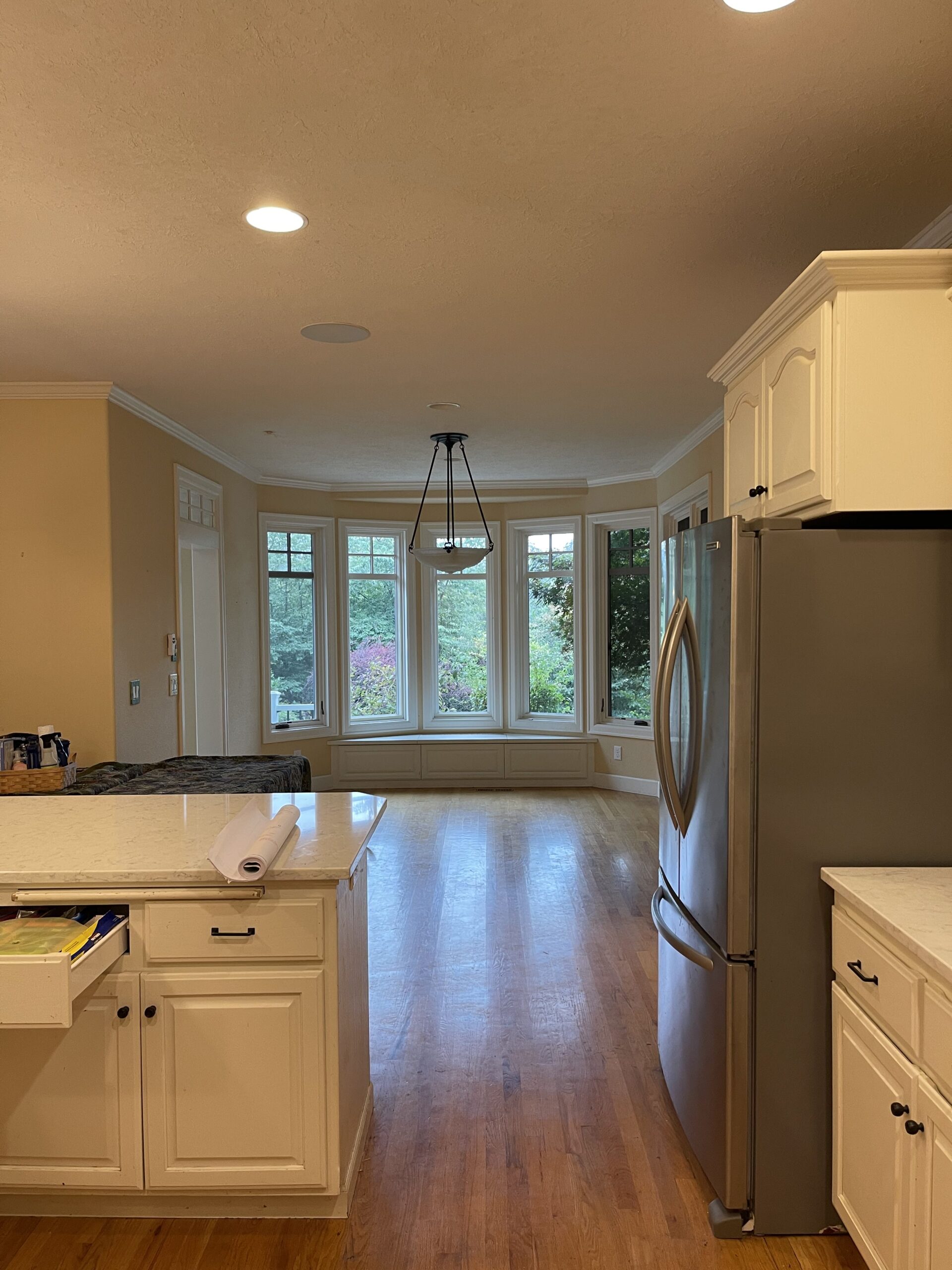 Old kitchen with white cabinets, stainless steel fridge, and large windows overlooking a lush green backyard.