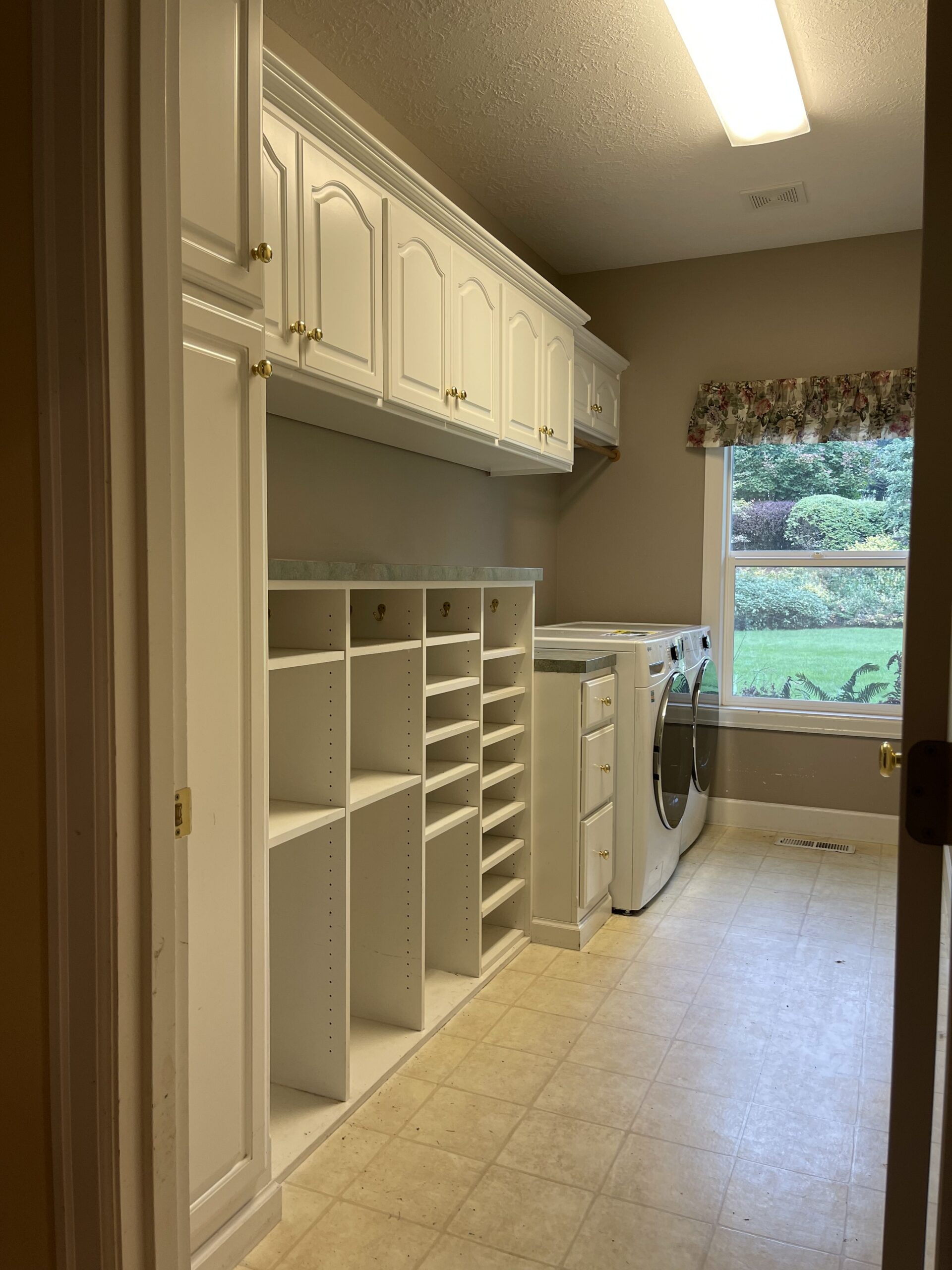 Old laundry room with white cabinets, shelves, and appliances near a window overlooking a garden.