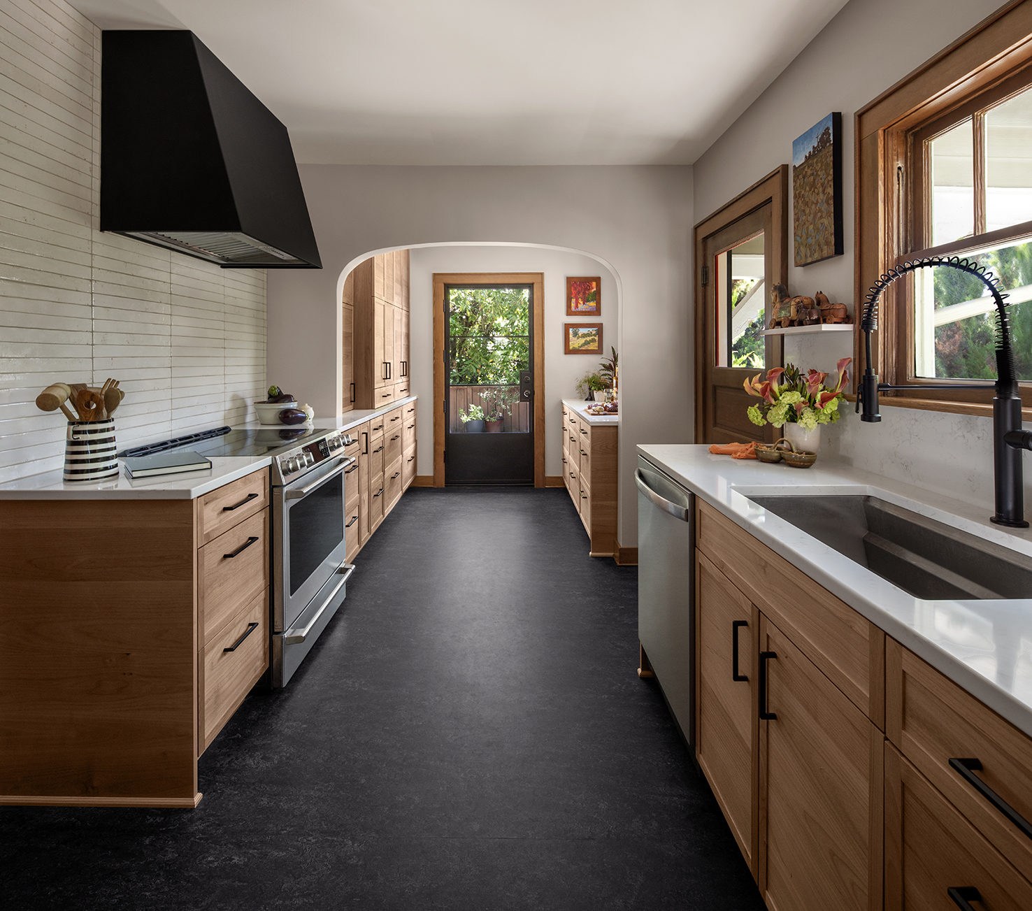 Modern kitchen with wooden cabinets, black range hood, and white countertops, leading to a garden-view door.