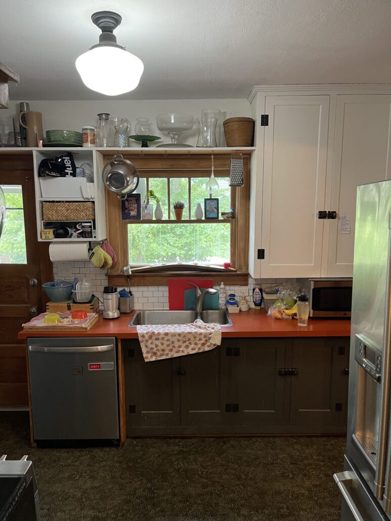 Cozy kitchen with red countertop, sink, and window. Shelves with dishes and decor. Brightly lit and organized space.