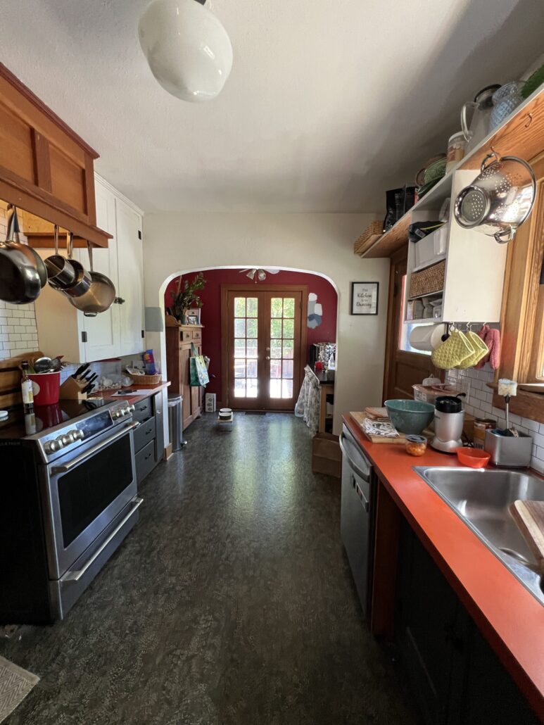 Cozy kitchen with red counters, hanging pots, and rustic decor leading to sunlit double doors.